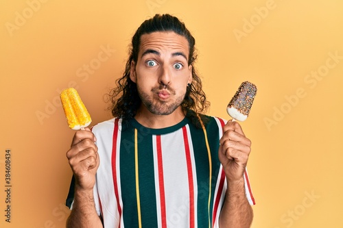 Young handsome man with long hair eating two ice cream puffing cheeks with funny face. mouth inflated with air, catching air. photo