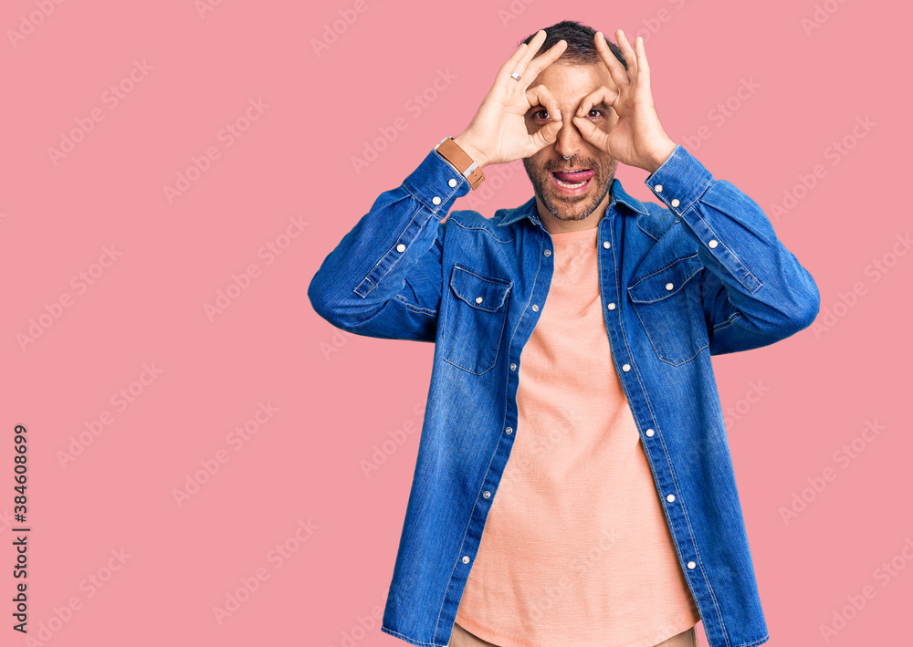 Young handsome man wearing casual clothes doing ok gesture like binoculars sticking tongue out, eyes looking through fingers. crazy expression. Young handsome man wearing casual clothes doing ok gesture like binoculars sticking tongue out, eyes looking through fingers. crazy expression.