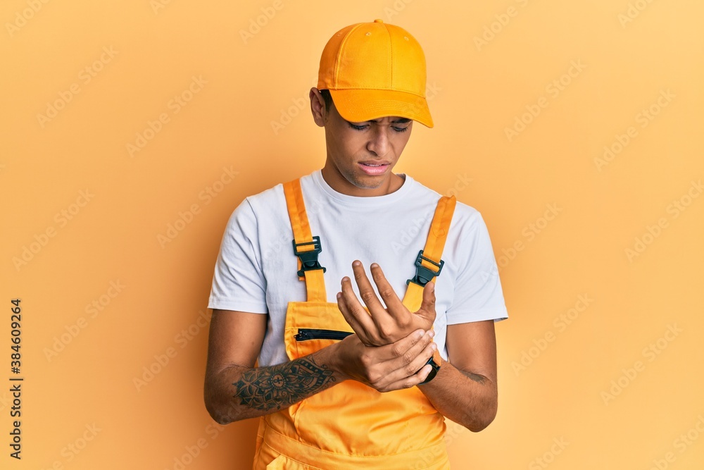 Young handsome african american man wearing handyman uniform over yellow background suffering pain on hands and fingers, arthritis inflammation Young handsome african american man wearing handyman uniform over yellow background suffering pain on hands and fingers, arthritis inflammation