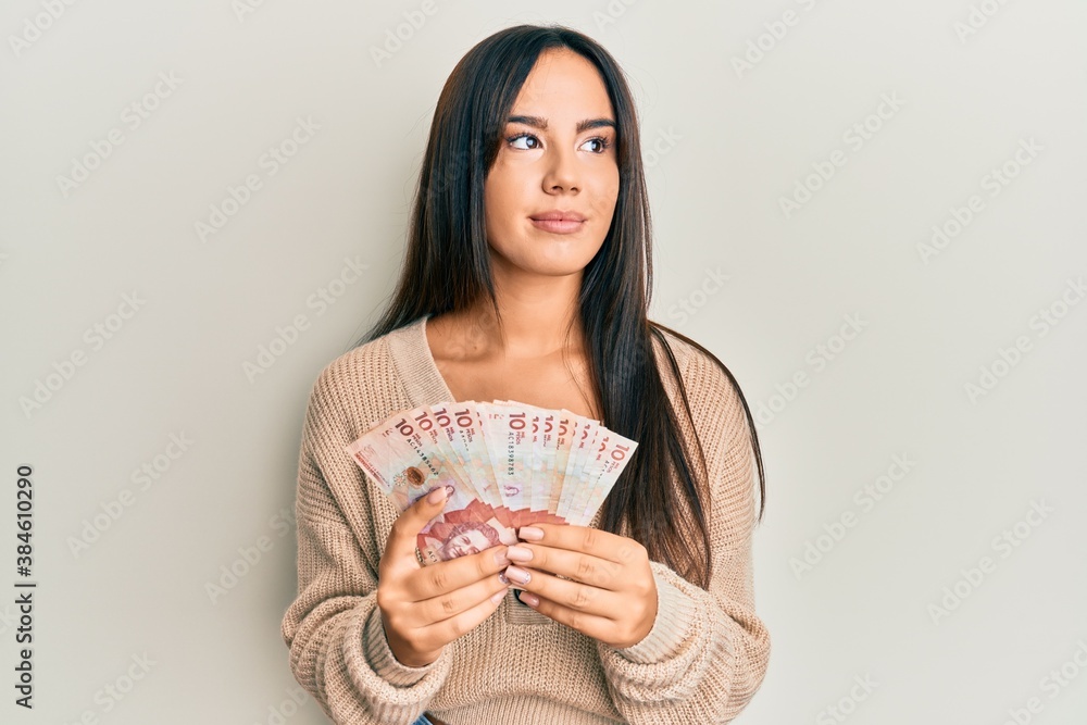 Young beautiful hispanic girl holding 10 colombian pesos banknotes smiling looking to the side and staring away thinking. Young beautiful hispanic girl holding 10 colombian pesos banknotes smiling looking to the side and staring away thinking.
