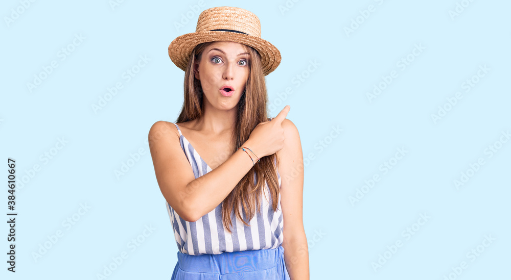 Young beautiful blonde woman wearing summer hat surprised pointing with finger to the side, open mouth amazed expression.