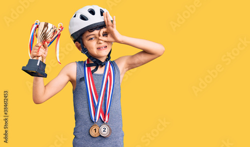 Little cute boy kid wearing bike helmet and winner medals holding winner trophy smiling happy doing ok sign with hand on eye looking through fingers