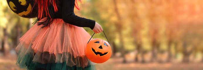 Little girl in witch costume with a balloon and a pumpkin bucket playing in autumn park. Child having fun at Halloween trick or treat. Kids trick or treating.