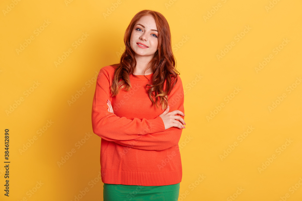 Portrait of young woman of Caucasian appearance wearing casual attire, posing at blank wall with arms crossed, feeling happy, smiles gently, red haired female in orange jumper. Portrait of young woman of Caucasian appearance wearing casual attire, posing at blank wall with arms crossed, feeling happy, smiles gently, red haired female in orange jumper.