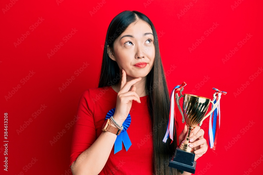 Young chinese woman wearing first place badge holding trophy serious face thinking about question with hand on chin, thoughtful about confusing idea Young chinese woman wearing first place badge holding trophy serious face thinking about question with hand on chin, thoughtful about confusing idea