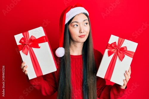 Young chinese woman wearing christmas hat and holding gifts smiling looking to the side and staring away thinking.