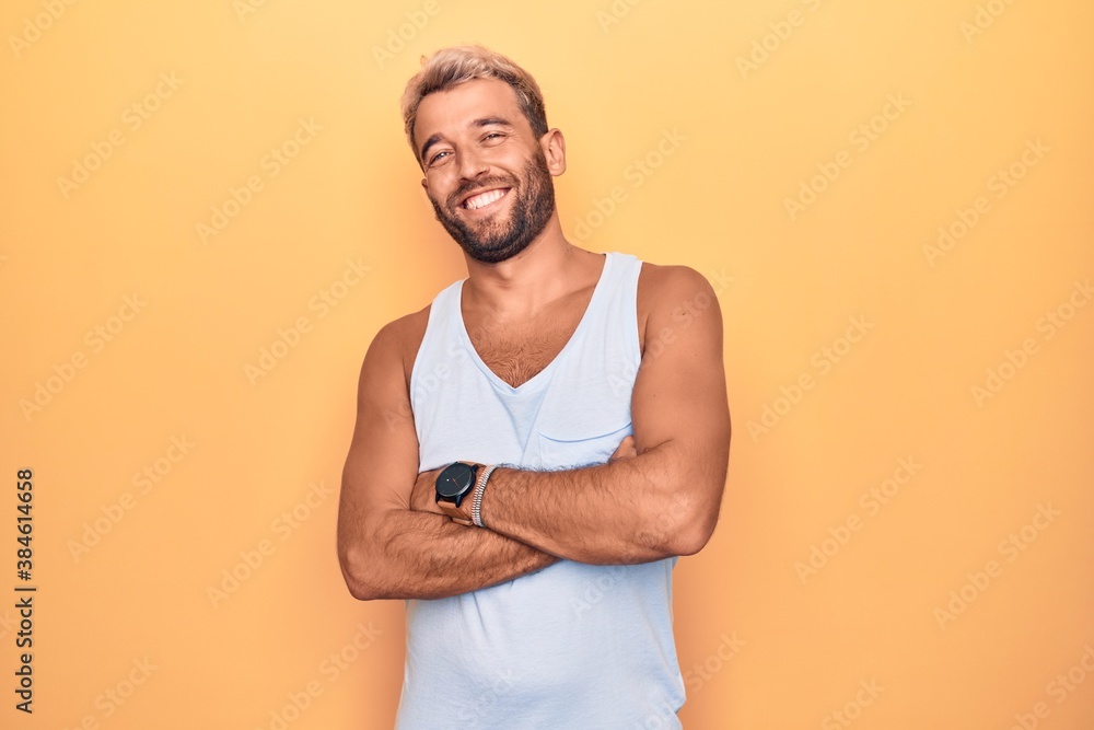 Young handsome blond man wearing casual sleeveless t-shirt over isolated yellow background happy face smiling with crossed arms looking at the camera. Positive person. Young handsome blond man wearing casual sleeveless t-shirt over isolated yellow background happy face smiling with crossed arms looking at the camera. Positive person.