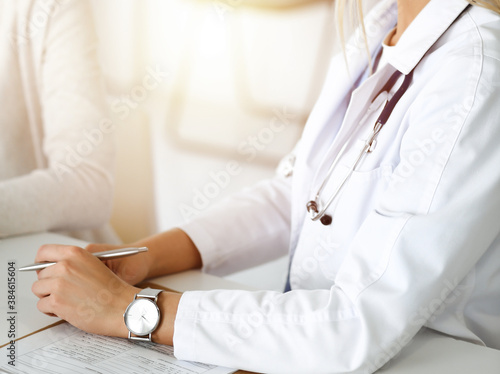 Unknown woman-doctor communicating with female patient in sunny clinic. Physician checks medical history record and exam results  close-up