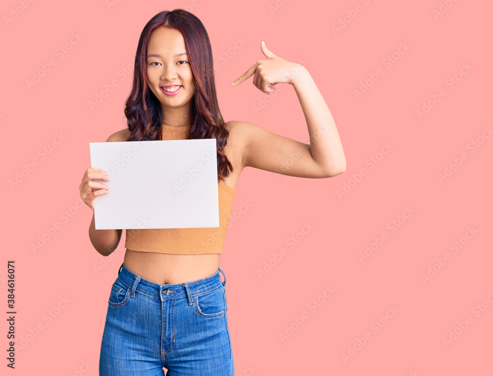 Young beautiful chinese girl holding paper banner with blank space pointing finger to one self smiling happy and proud Young beautiful chinese girl holding paper banner with blank space pointing finger to one self smiling happy and proud