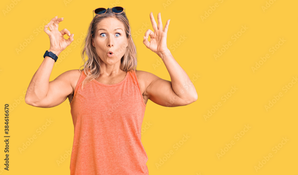 Middle age fit blonde woman wearing casual summer clothes and sunglasses looking surprised and shocked doing ok approval symbol with fingers. crazy expression