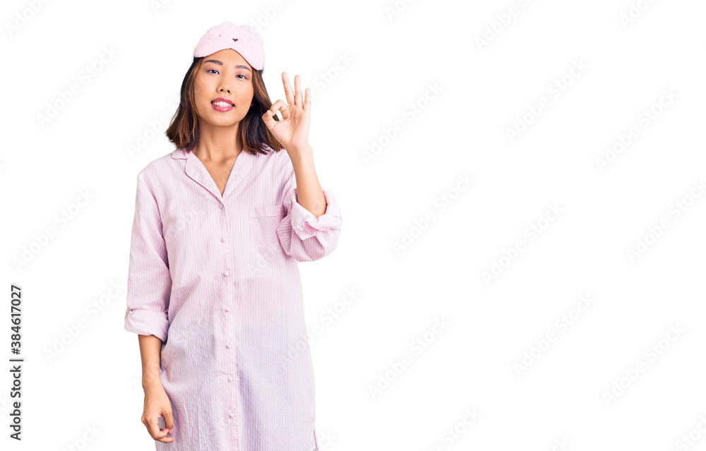 Young beautiful chinese girl wearing sleep mask and pajama smiling positive doing ok sign with hand and fingers. successful expression. Young beautiful chinese girl wearing sleep mask and pajama smiling positive doing ok sign with hand and fingers. successful expression.