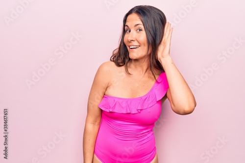 Young beautiful brunette woman wearing pink swimwear standing over isolated background smiling with hand over ear listening and hearing to rumor or gossip. Deafness concept.