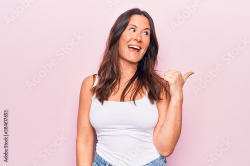 Young beautiful brunette woman wearing casual sleeveless t-shirt over white background pointing thumb up to the side smiling happy with open mouth © Krakenimages.com