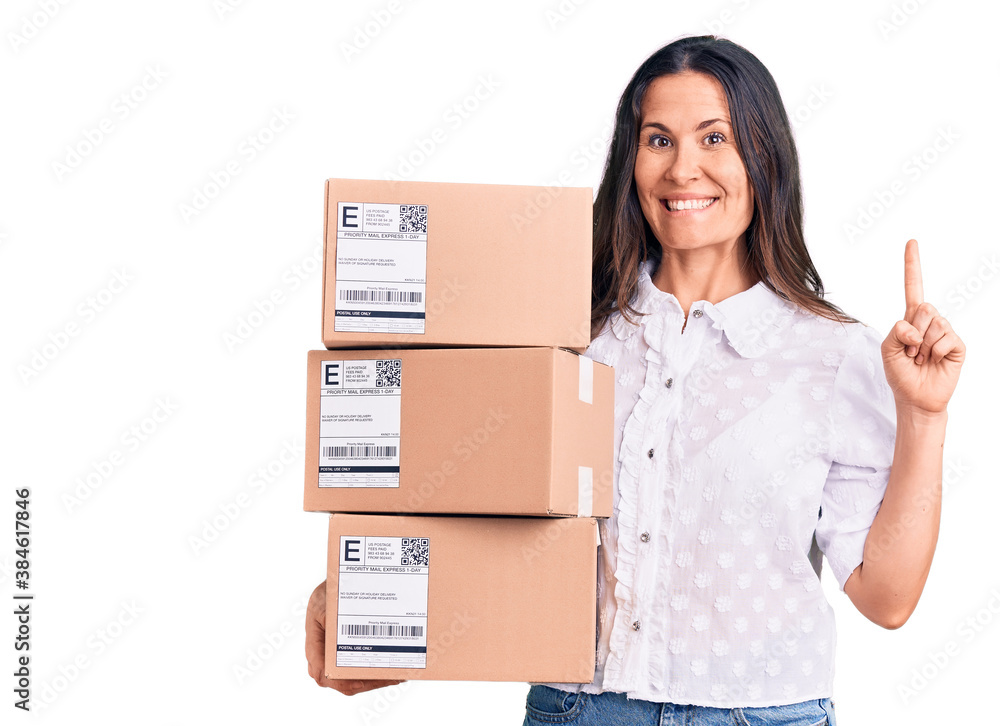 Young beautiful brunette woman holding delivery cardboard package surprised with an idea or question pointing finger with happy face, number one Young beautiful brunette woman holding delivery cardboard package surprised with an idea or question pointing finger with happy face, number one