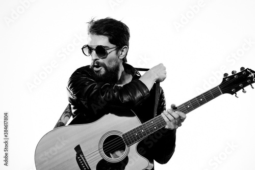 A man plays the guitar in a black leather jacket with sunglasses on a light background
