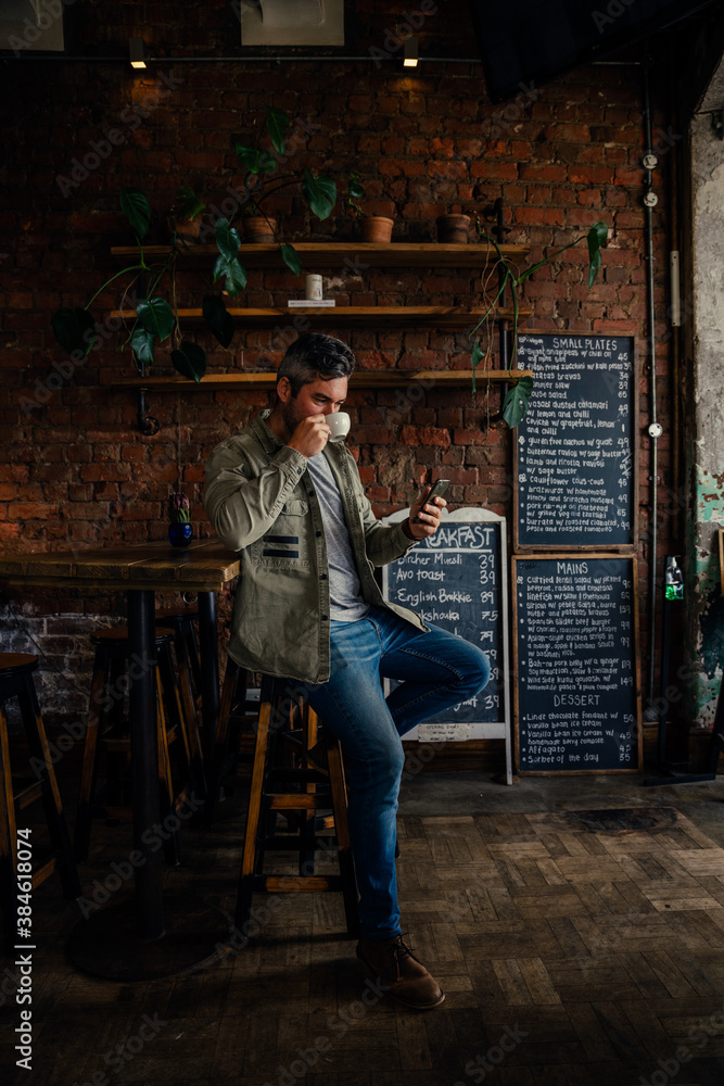 Fototapeta premium Male customer sitting in authentic cafe sipping hot coffee chatting to mates on smartphone