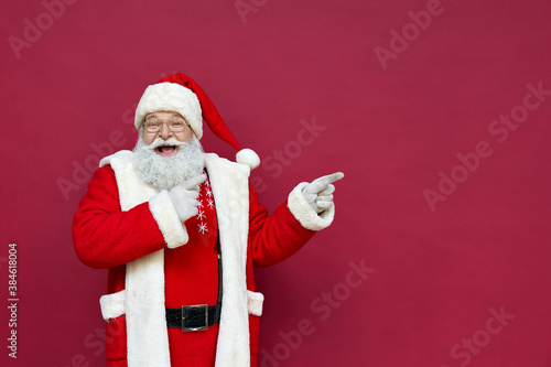 Funny happy excited old bearded Santa Claus face wearing costume looking at camera showing pointing fingers aside advertising Christmas promotion, New Year xmas discount ad isolated on red background. photo