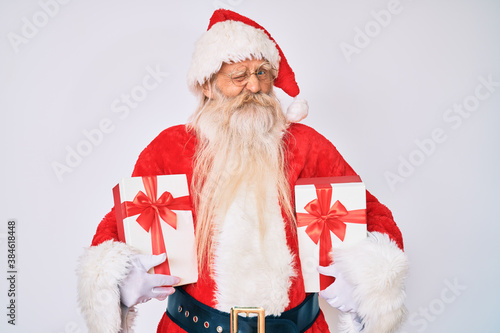 Fotografia Old senior man wearing santa claus costume holding presents winking looking at the camera with sexy expression, cheerful and happy face