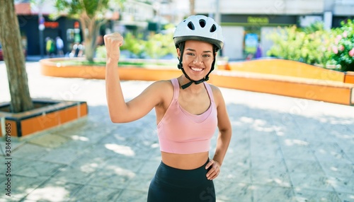 Young beautiful hispanic cyclist woman smiling happy wearing bike helmet at the town with raising arm doing strong gesture