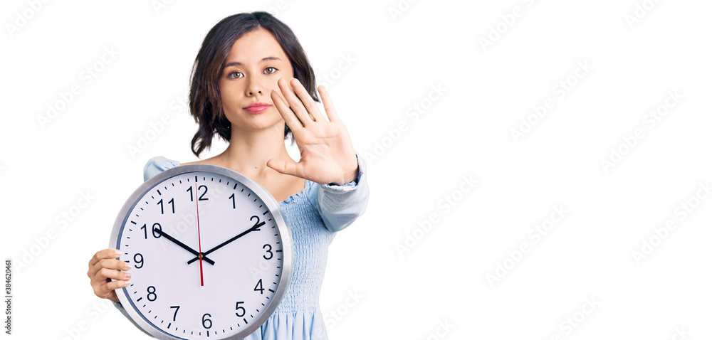Young beautiful girl holding big clock with open hand doing stop sign with serious and confident expression, defense gesture Young beautiful girl holding big clock with open hand doing stop sign with serious and confident expression, defense gesture