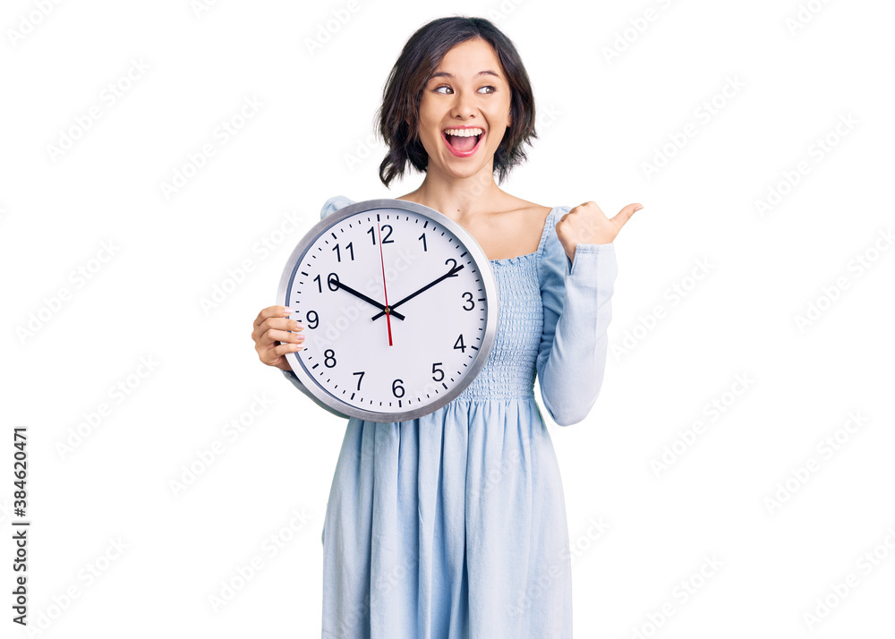Young beautiful girl holding big clock pointing thumb up to the side smiling happy with open mouth Young beautiful girl holding big clock pointing thumb up to the side smiling happy with open mouth