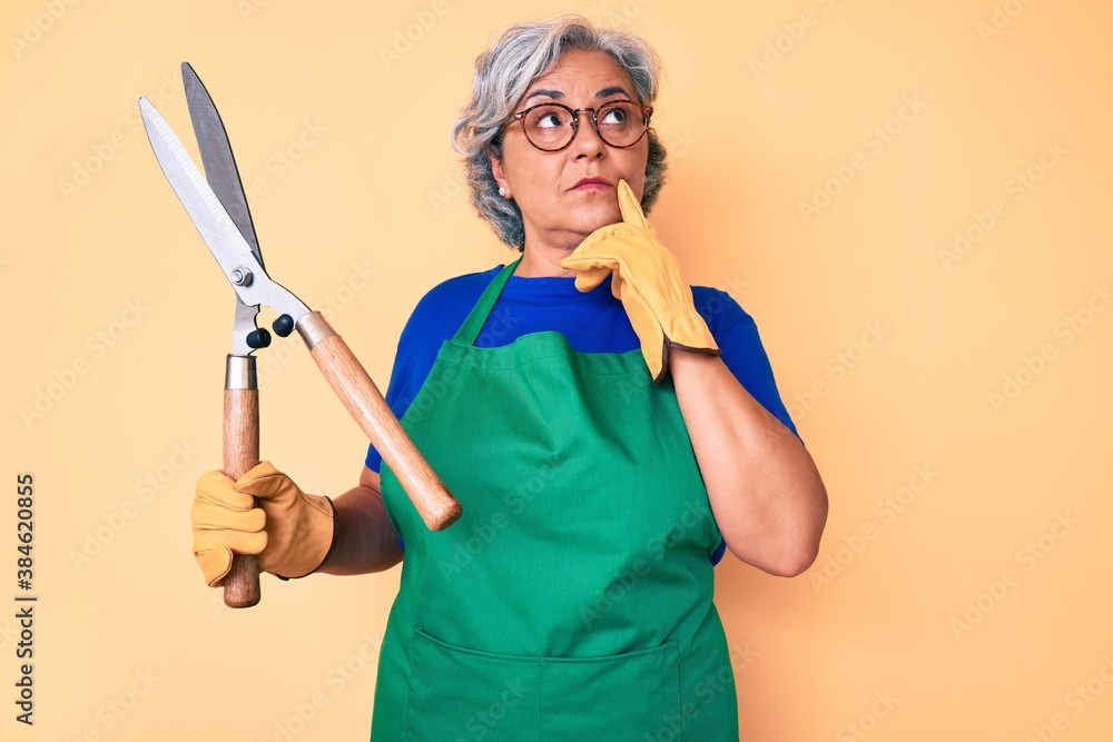Senior hispanic woman wearing gardener apron and gloves holding shears serious face thinking about question with hand on chin, thoughtful about confusing idea Senior hispanic woman wearing gardener apron and gloves holding shears serious face thinking about question with hand on chin, thoughtful about confusing idea