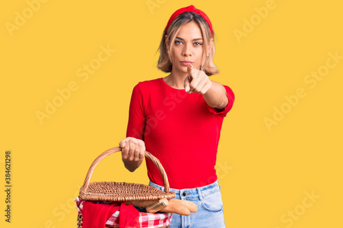 Beautiful caucasian woman holding picnic wicker basket with bread pointing with finger to the camera and to you, confident gesture looking serious