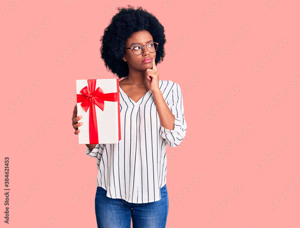 Young african american woman holding gift serious face thinking about question with hand on chin, thoughtful about confusing idea Young african american woman holding gift serious face thinking about question with hand on chin, thoughtful about confusing idea