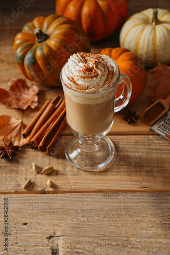 Spice pumpkin latte with cream foam, cinnamon stick, leaf and orange pumpkins on wooden background