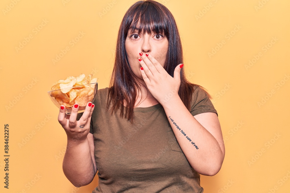 Young plus size woman holding potato chip covering mouth with hand, shocked and afraid for mistake. surprised expression