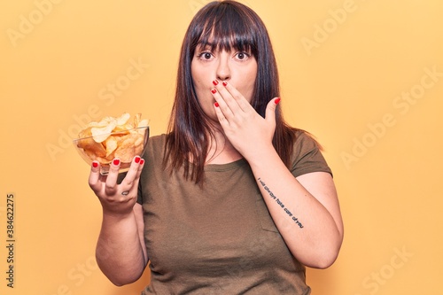 Young plus size woman holding potato chip covering mouth with hand  shocked and afraid for mistake. surprised expression