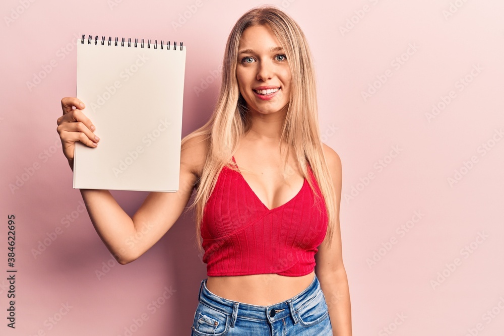 Young blonde woman holding notebook looking positive and happy standing and smiling with a confident smile showing teeth Young blonde woman holding notebook looking positive and happy standing and smiling with a confident smile showing teeth