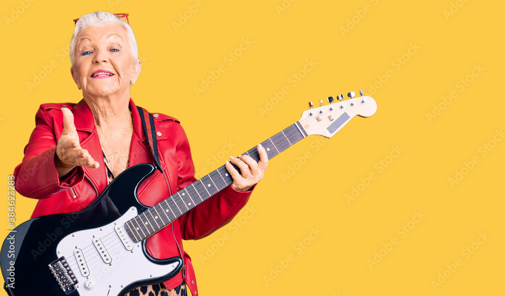 Fototapeta premium Senior beautiful woman with blue eyes and grey hair wearing a modern look playing electric guitar smiling friendly offering handshake as greeting and welcoming. successful business.