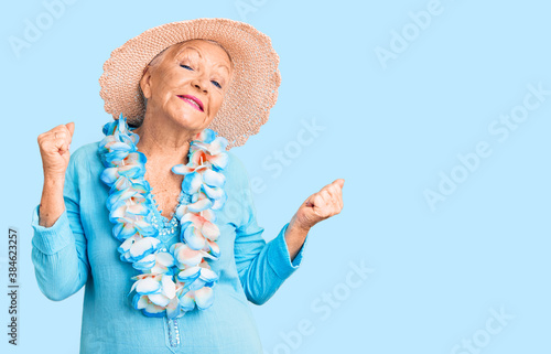 Senior beautiful woman with blue eyes and grey hair wearing summer hat and hawaiian lei very happy and excited doing winner gesture with arms raised, smiling and screaming for success. celebration. © Krakenimages.com