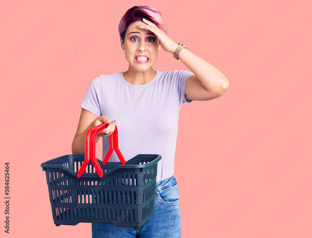 Young beautiful woman with pink hair holding supermarket shopping basket stressed and frustrated with hand on head, surprised and angry face