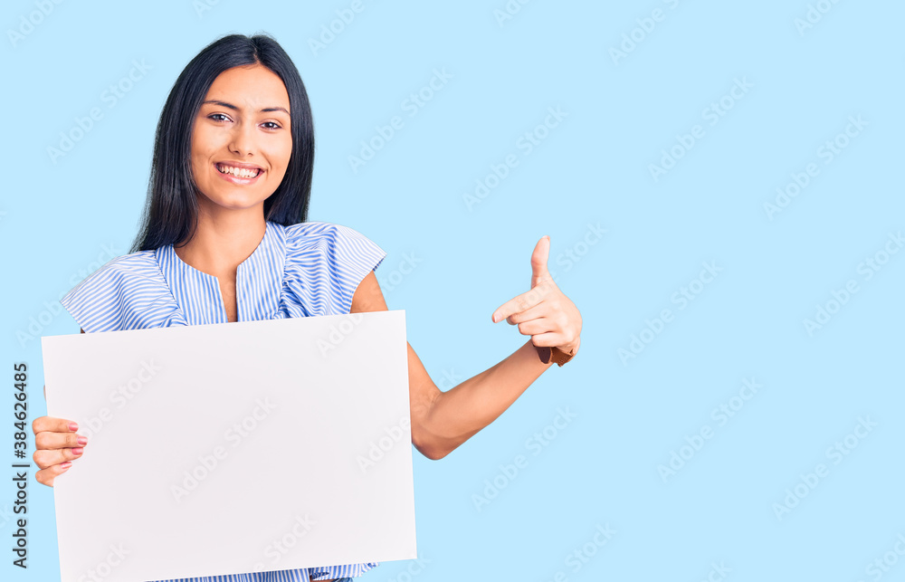 Young beautiful latin girl holding blank empty banner smiling happy pointing with hand and finger Young beautiful latin girl holding blank empty banner smiling happy pointing with hand and finger
