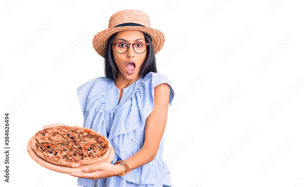 Young beautiful latin girl wearing summer hat and glasses holding pizza scared and amazed with open mouth for surprise, disbelief face Young beautiful latin girl wearing summer hat and glasses holding pizza scared and amazed with open mouth for surprise, disbelief face