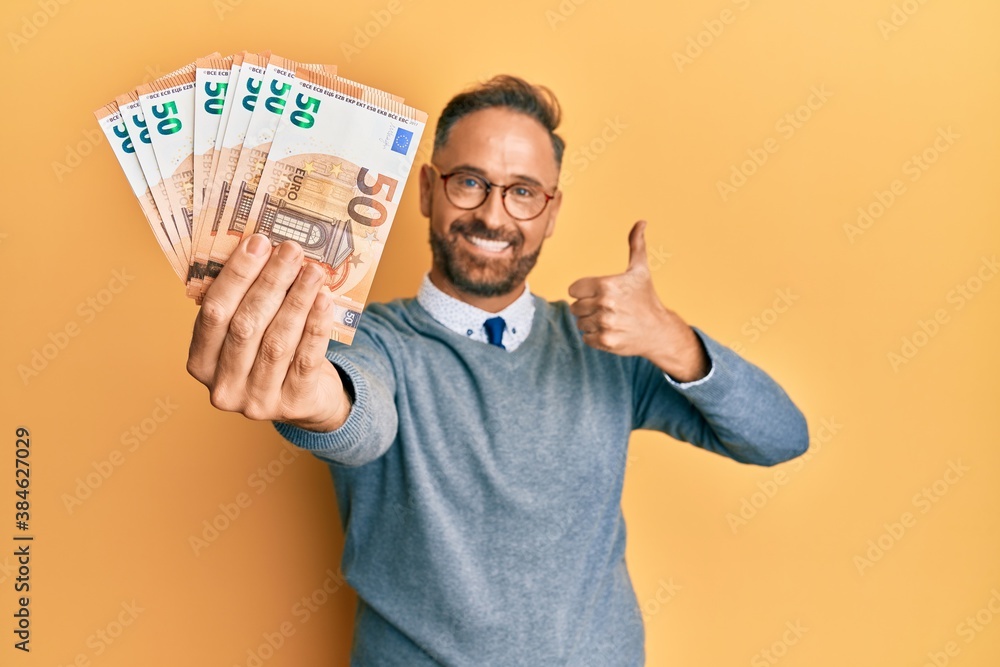 Handsome middle age man holding bunch of euros smiling happy and positive, thumb up doing excellent and approval sign Handsome middle age man holding bunch of euros smiling happy and positive, thumb up doing excellent and approval sign