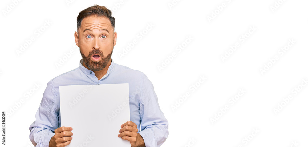 Handsome middle age man holding blank empty banner scared and amazed with open mouth for surprise, disbelief face Handsome middle age man holding blank empty banner scared and amazed with open mouth for surprise, disbelief face