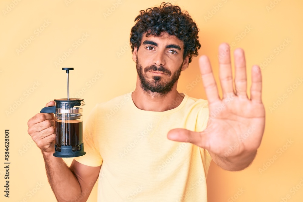Handsome young man with curly hair and bear holding french coffee maker with open hand doing stop sign with serious and confident expression, defense gesture Handsome young man with curly hair and bear holding french coffee maker with open hand doing stop sign with serious and confident expression, defense gesture