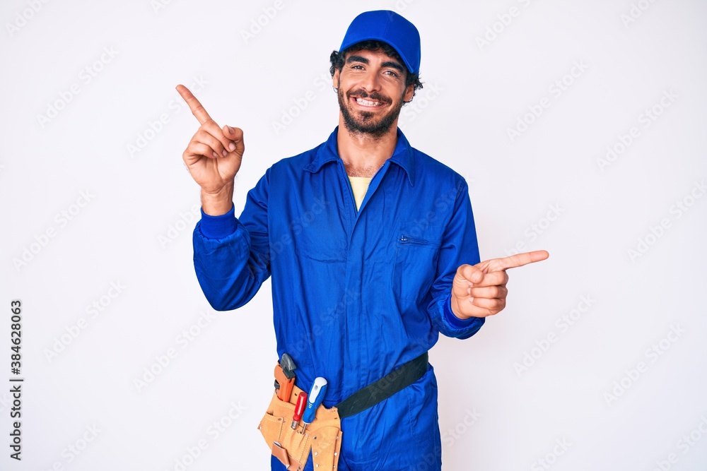 Handsome young man with curly hair and bear weaing handyman uniform smiling confident pointing with fingers to different directions. copy space for advertisement