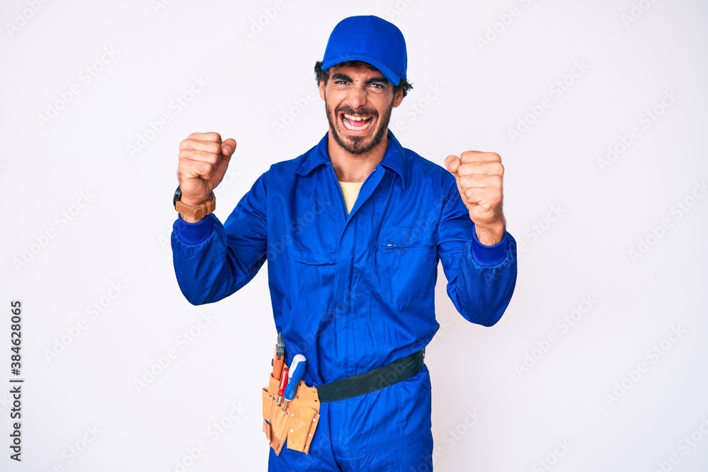 Handsome young man with curly hair and bear weaing handyman uniform angry and mad raising fists frustrated and furious while shouting with anger. rage and aggressive concept.