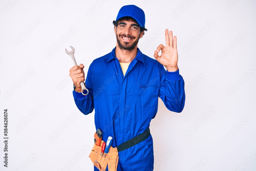 Handsome young man with curly hair and bear wearing builder jumpsuit uniform and holding wrench doing ok sign with fingers, smiling friendly gesturing excellent symbol