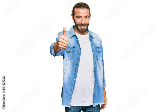 Attractive man with long hair and beard wearing casual denim jacket doing happy thumbs up gesture with hand. approving expression looking at the camera showing success.