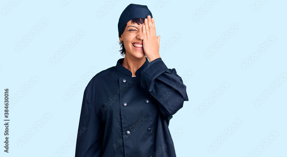 Young brunette woman with short hair wearing professional cook uniform covering one eye with hand, confident smile on face and surprise emotion. Young brunette woman with short hair wearing professional cook uniform covering one eye with hand, confident smile on face and surprise emotion.