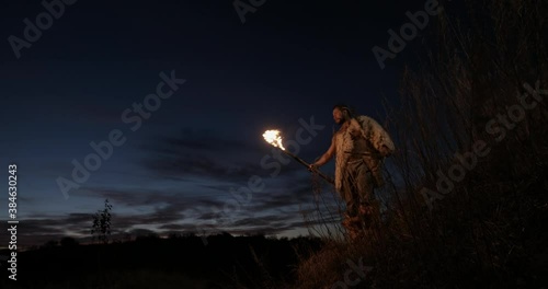 Primitive hunter gatherer wearing sheepskins and carrying a bow and arrows walking by torch light using a burning stick taper at night in countryside. Portrait of primeval caveman wearing animal night
