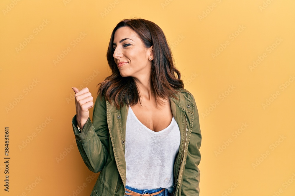 Young brunette woman wearing casual clothes over yellow background smiling with happy face looking and pointing to the side with thumb up. Young brunette woman wearing casual clothes over yellow background smiling with happy face looking and pointing to the side with thumb up.