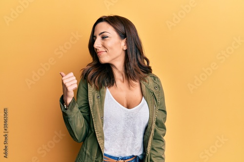 Young brunette woman wearing casual clothes over yellow background smiling with happy face looking and pointing to the side with thumb up.