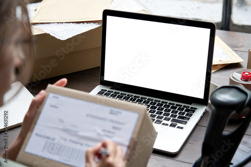 Female seller, internet shop small business owner holding ecommerce shipping box checking online store dropshipping order on mock up white blank laptop screen. E commerce post shipping delivery.