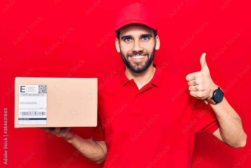 Young handsome man with beard wearing delivery uniform holding package smiling happy and positive, thumb up doing excellent and approval sign Young handsome man with beard wearing delivery uniform holding package smiling happy and positive, thumb up doing excellent and approval sign